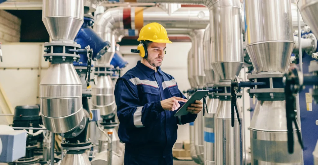 Picture of an engineer wearing a helmet and work uniform, holding a tablet, in the middle of an industrial place