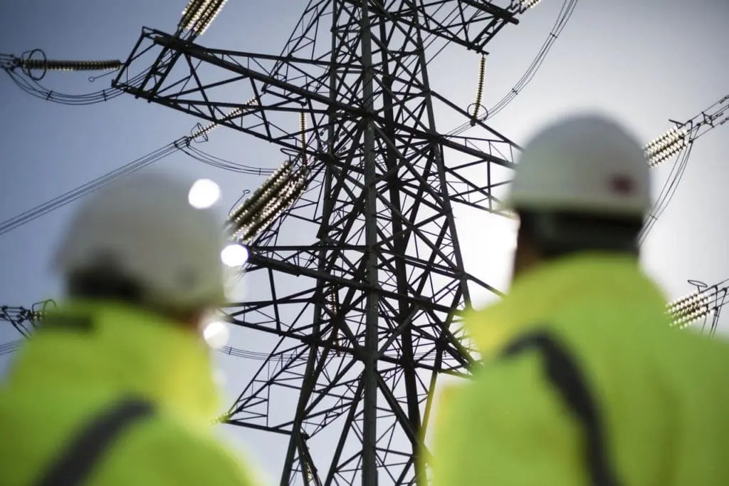 Two men wearing yellow vests and helmets looking at the electricity grid
